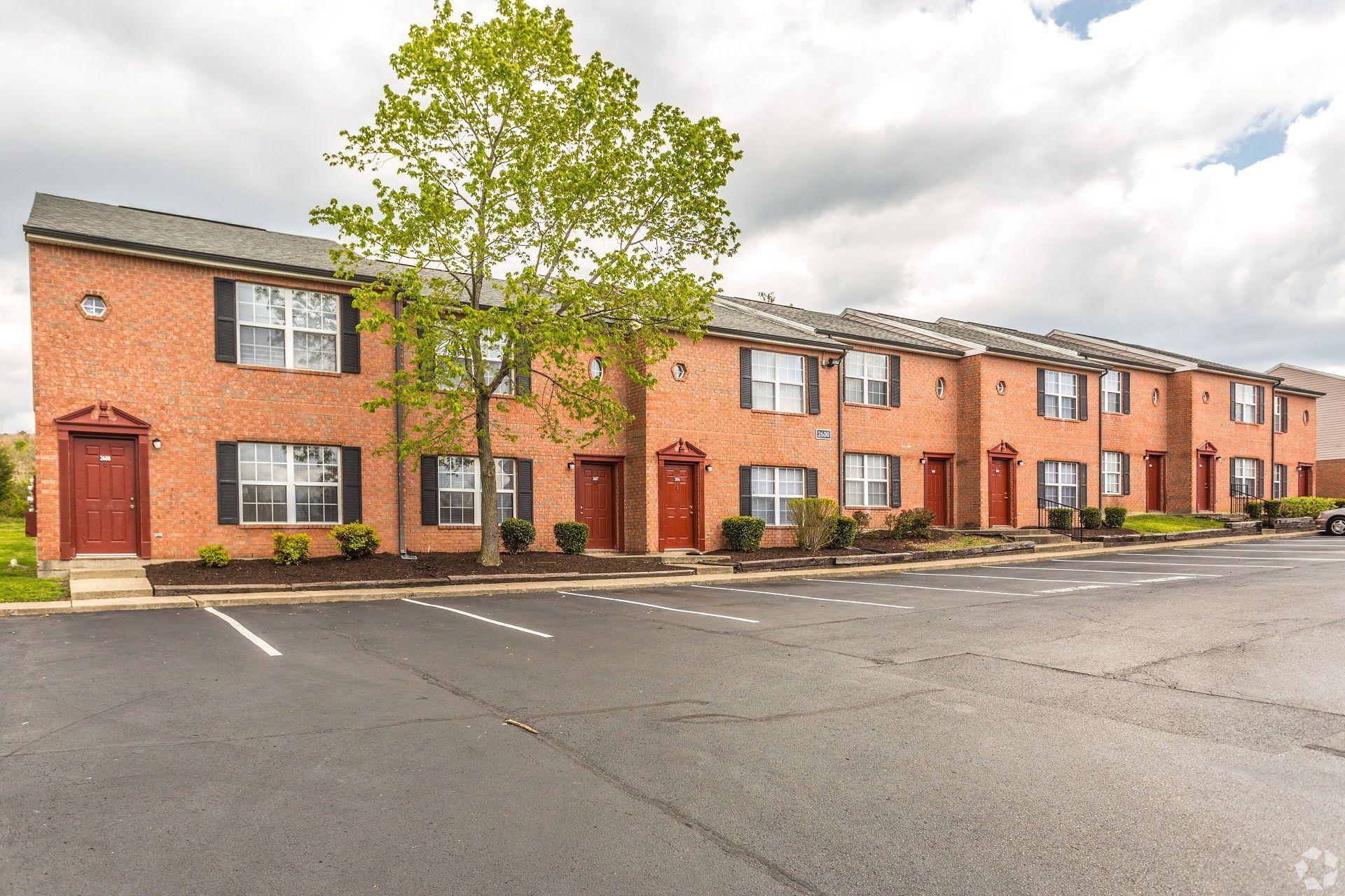 A two-story red brick apartment complex with several brown entry doors, white-trimmed windows, and a paved parking lot.