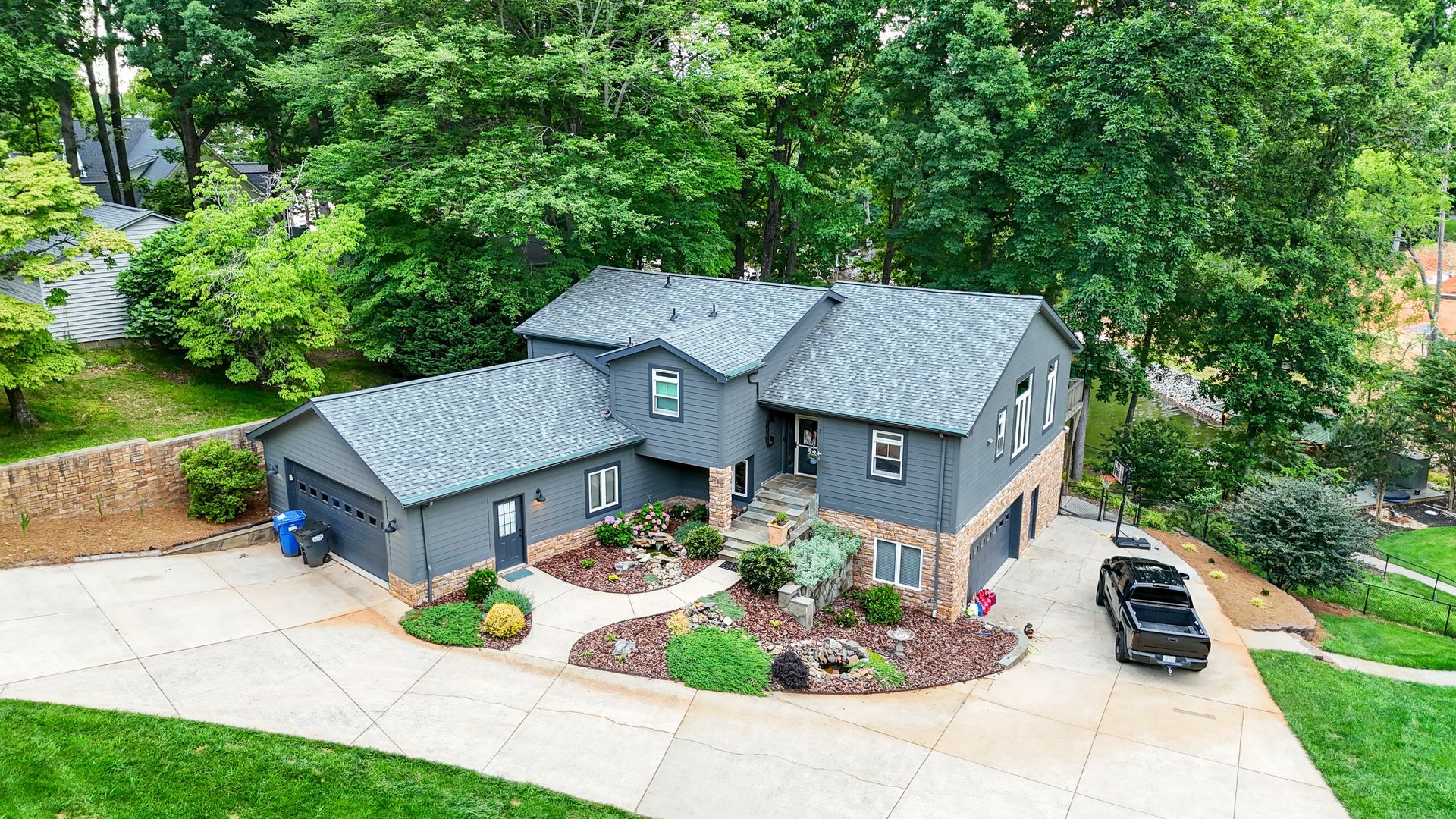 Gray house with stone and dark trim, surrounded by trees and greenery. A black truck is parked in the driveway.
