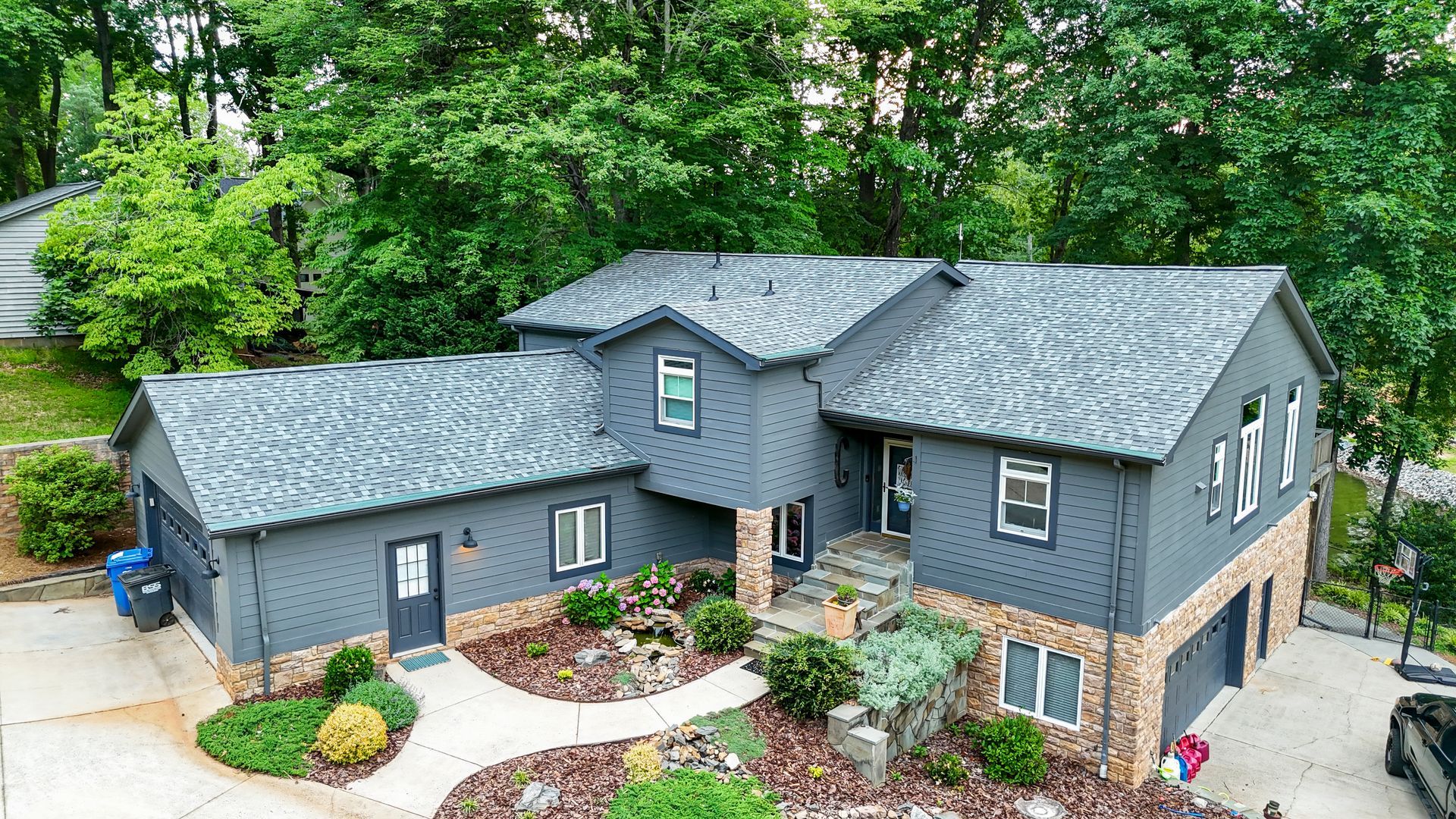 Gray two-story house with stone accents, surrounded by greenery and trees; driveway on the right.