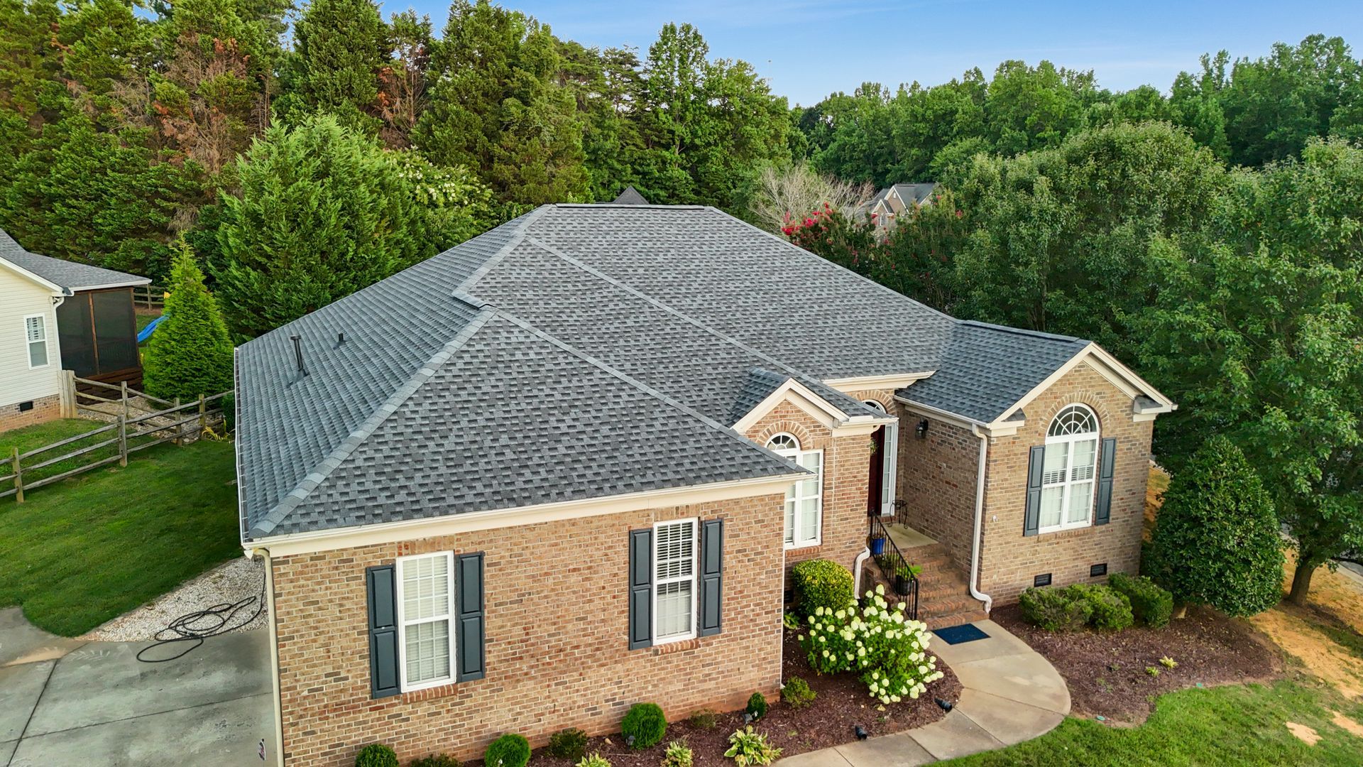 Brick house with dark roof and shutters, surrounded by green trees and lawn.