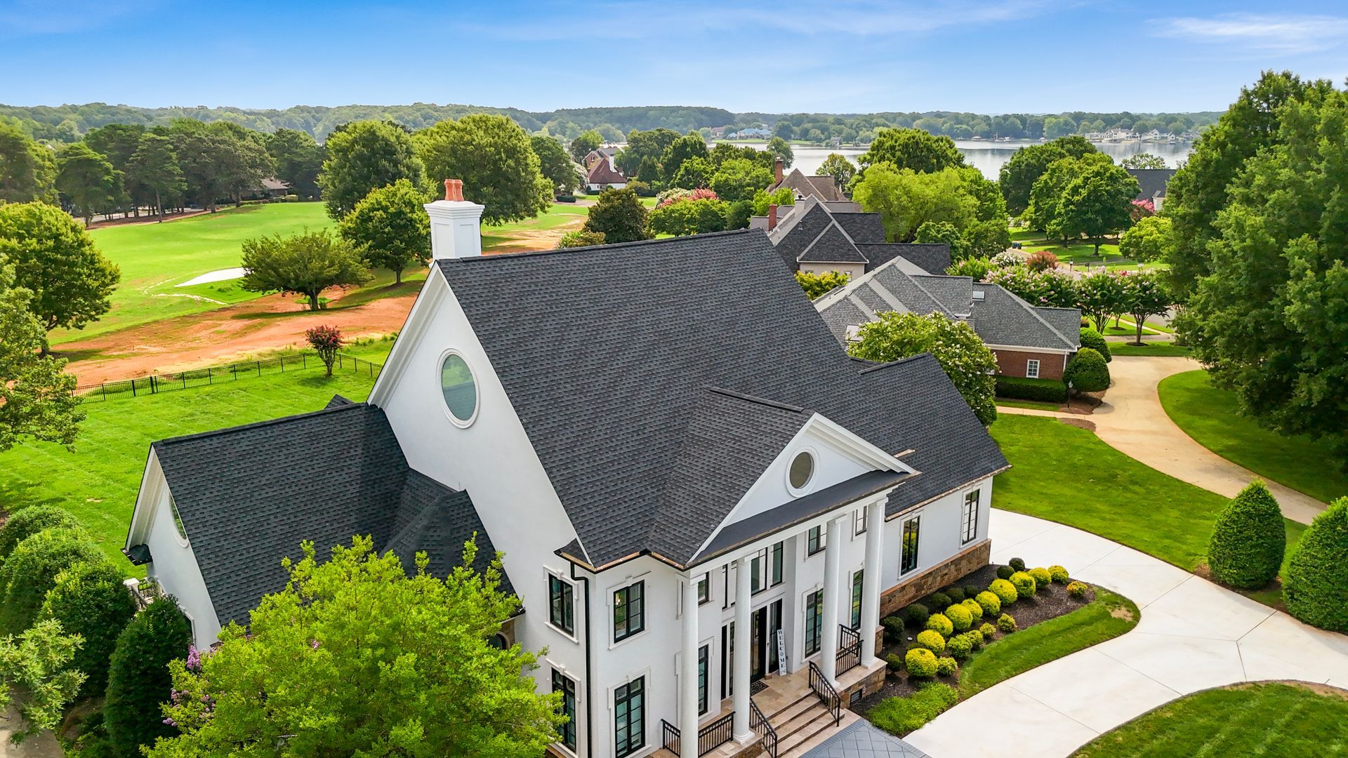 White mansion with dark roof and columns, on a green lawn, near trees and water.