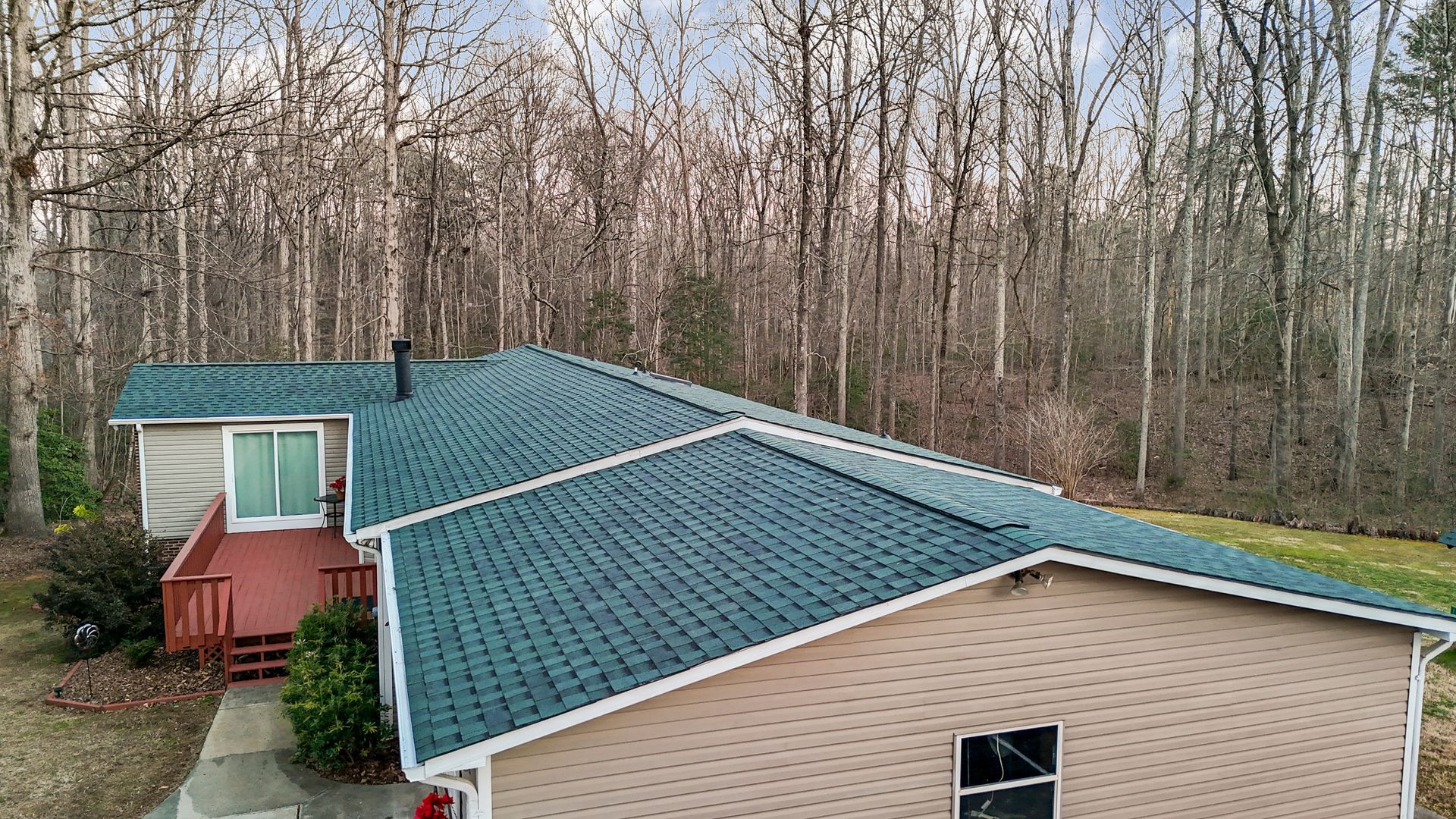 Tan house with a green roof and red deck, backed by a forest.