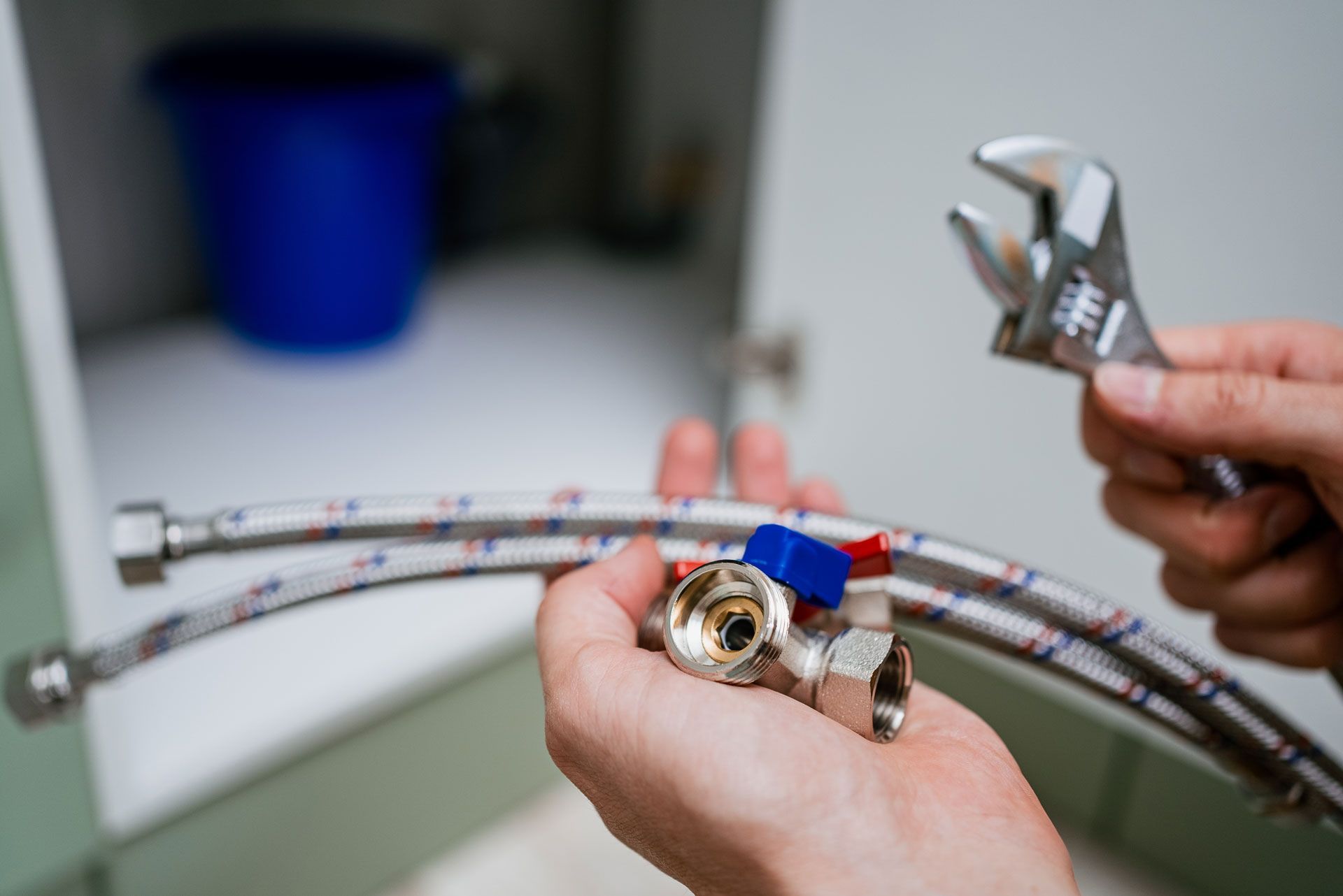 Hands holding braided hoses and a wrench while adjusting a plumbing valve.
