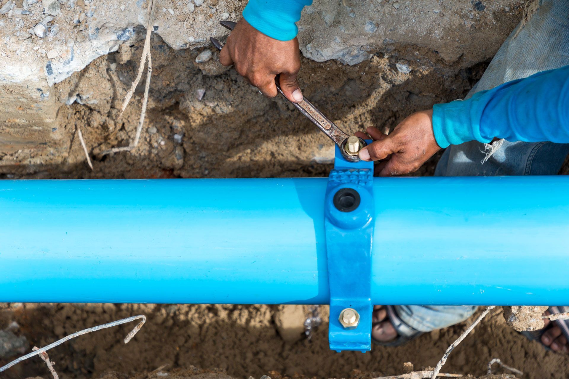 Hands tightening a clamp on a bright blue underground pipe in a dirt trench