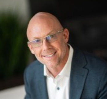 Bald man wearing glasses, white shirt, and blue suit jacket, smiling, indoor setting.