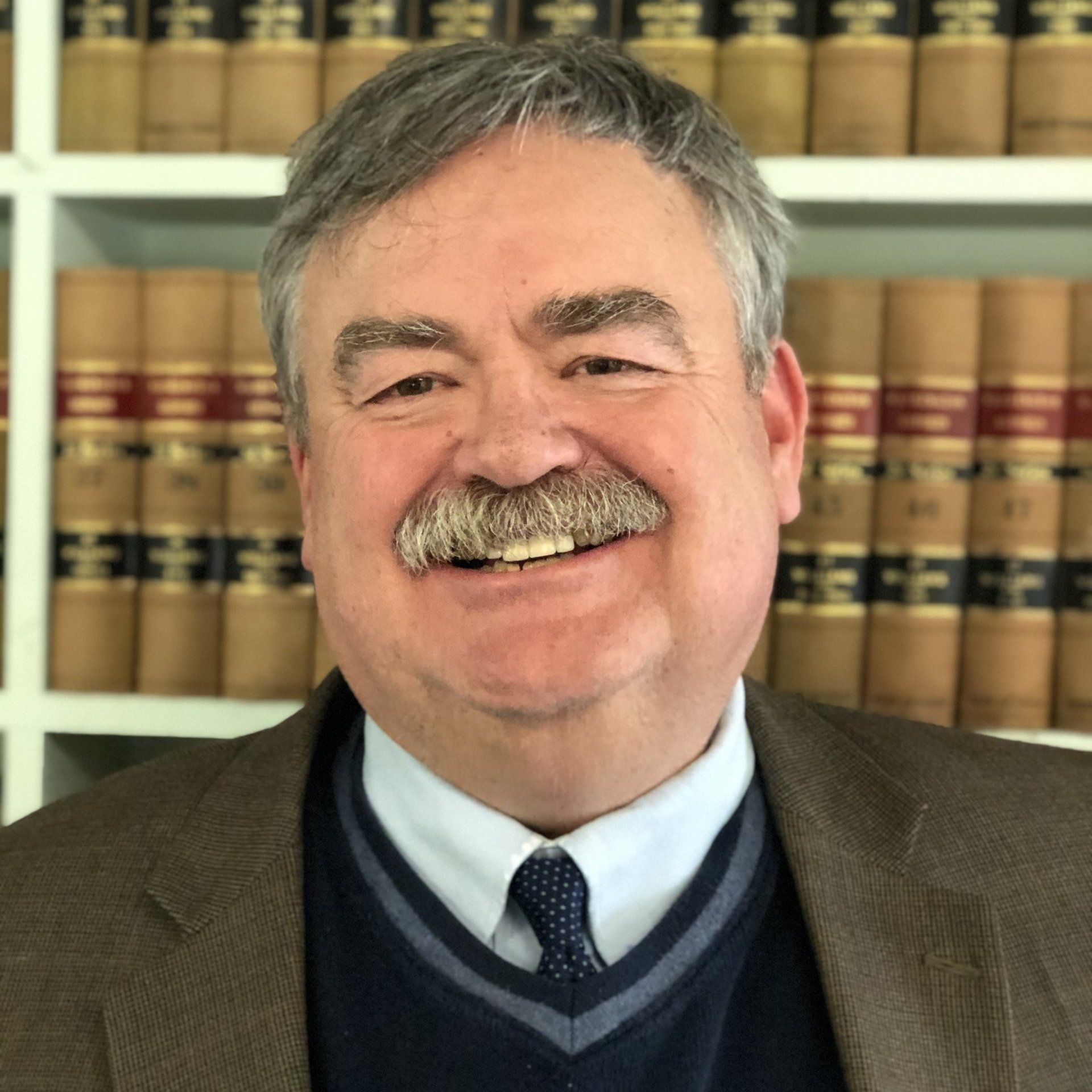 Man with mustache smiling, wearing a blue sweater and a brown jacket, in front of bookshelves with law books.