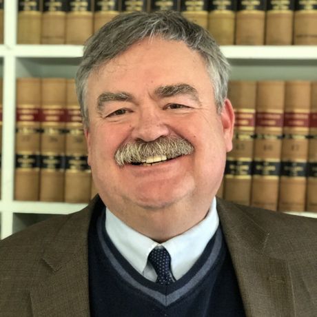Man with mustache smiling, wearing a blue sweater and a brown jacket, in front of bookshelves with law books.