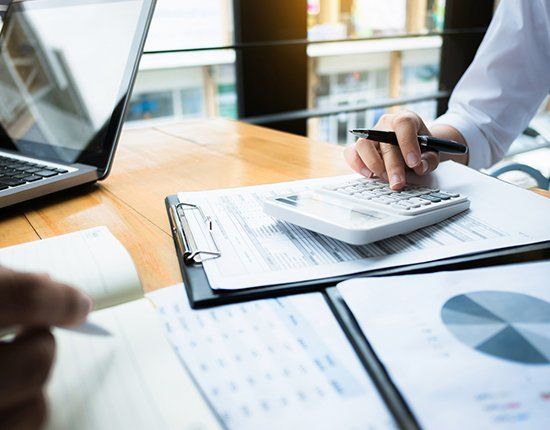Person calculating on a calculator over documents on a wooden desk, near a laptop and pie chart.