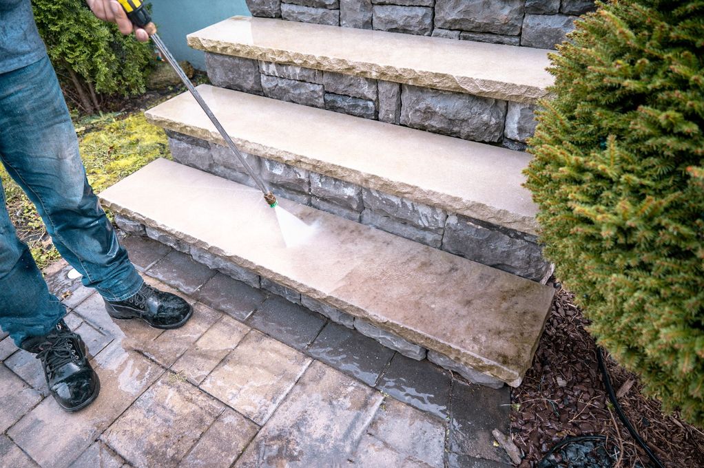 A person uses a pressure washer to clean stone steps outdoors, showing the contrast between dirty and clean sections.