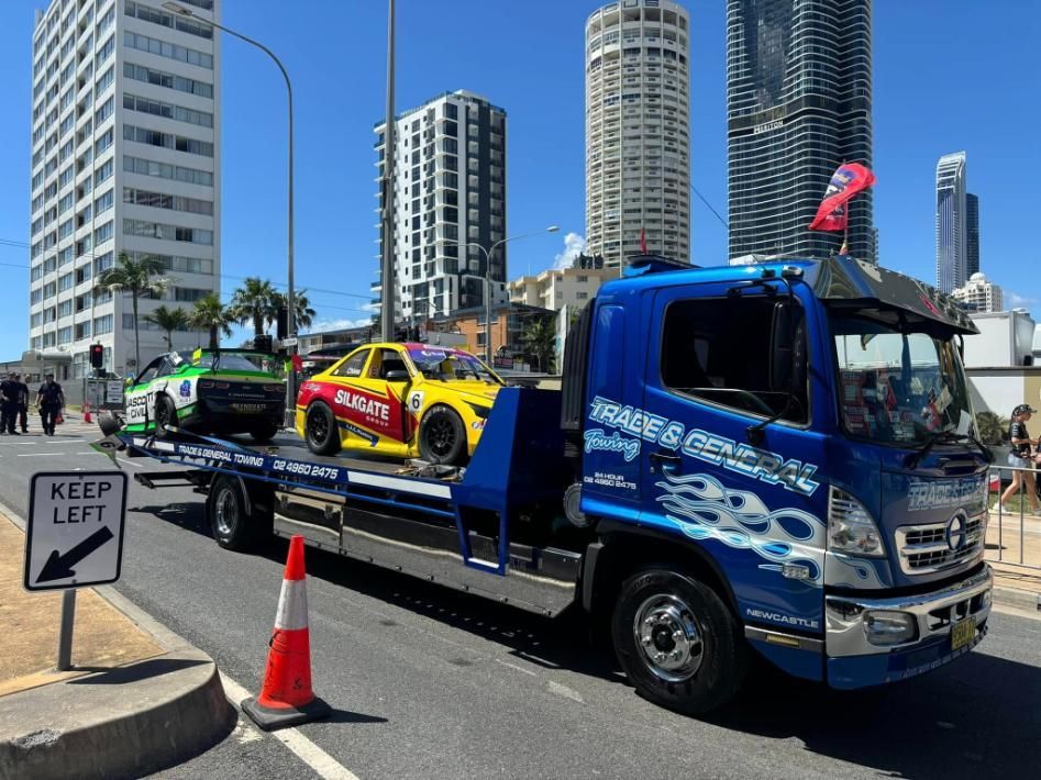 A Blue Tow Truck With Two Race Cars on the Back is Parked on the Side of the Road — Trade & General Towing in Cessnock, NSW