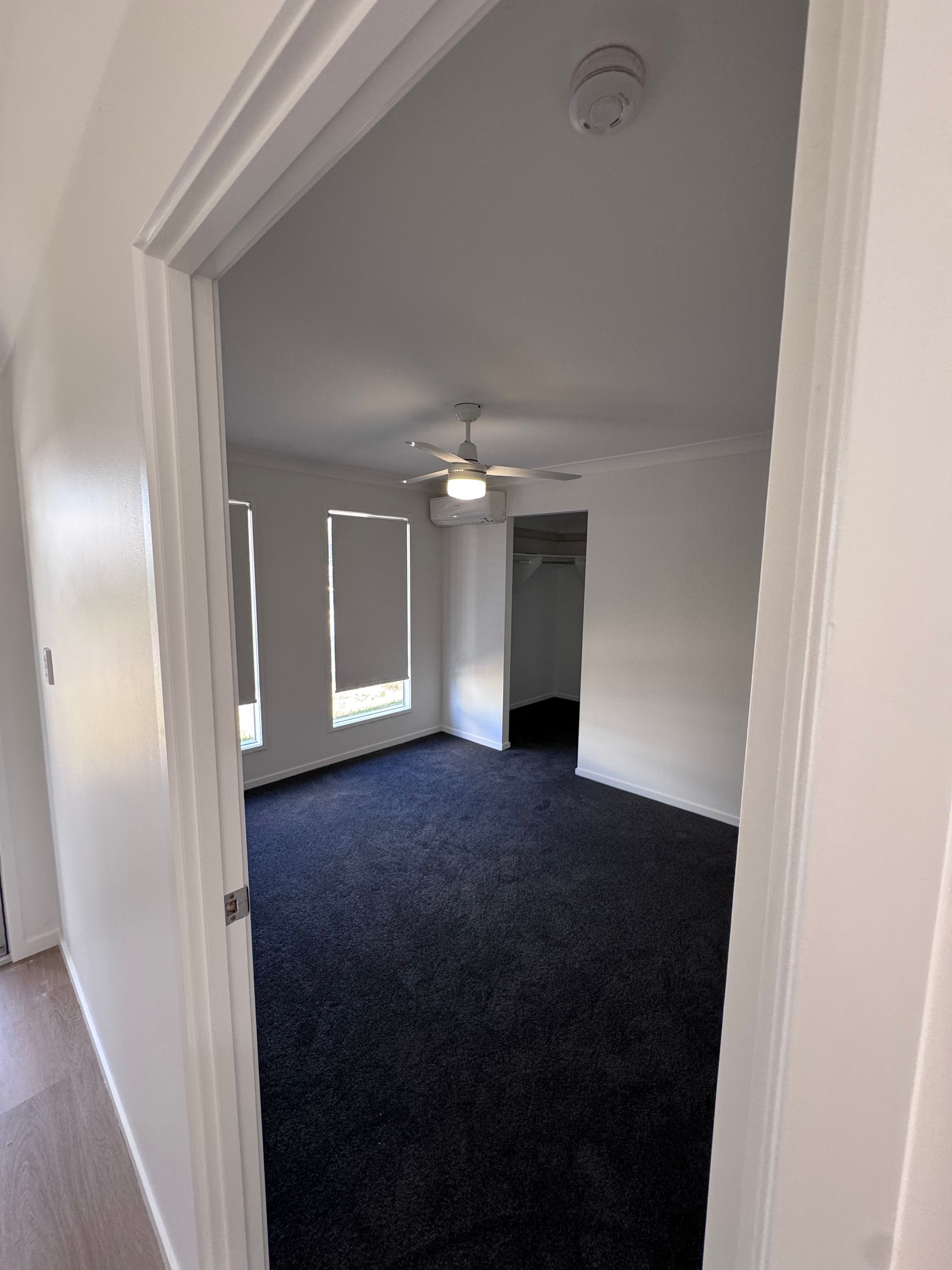 Empty carpeted room viewed through a doorway, with white walls, ceiling fan, and daylight from windows.