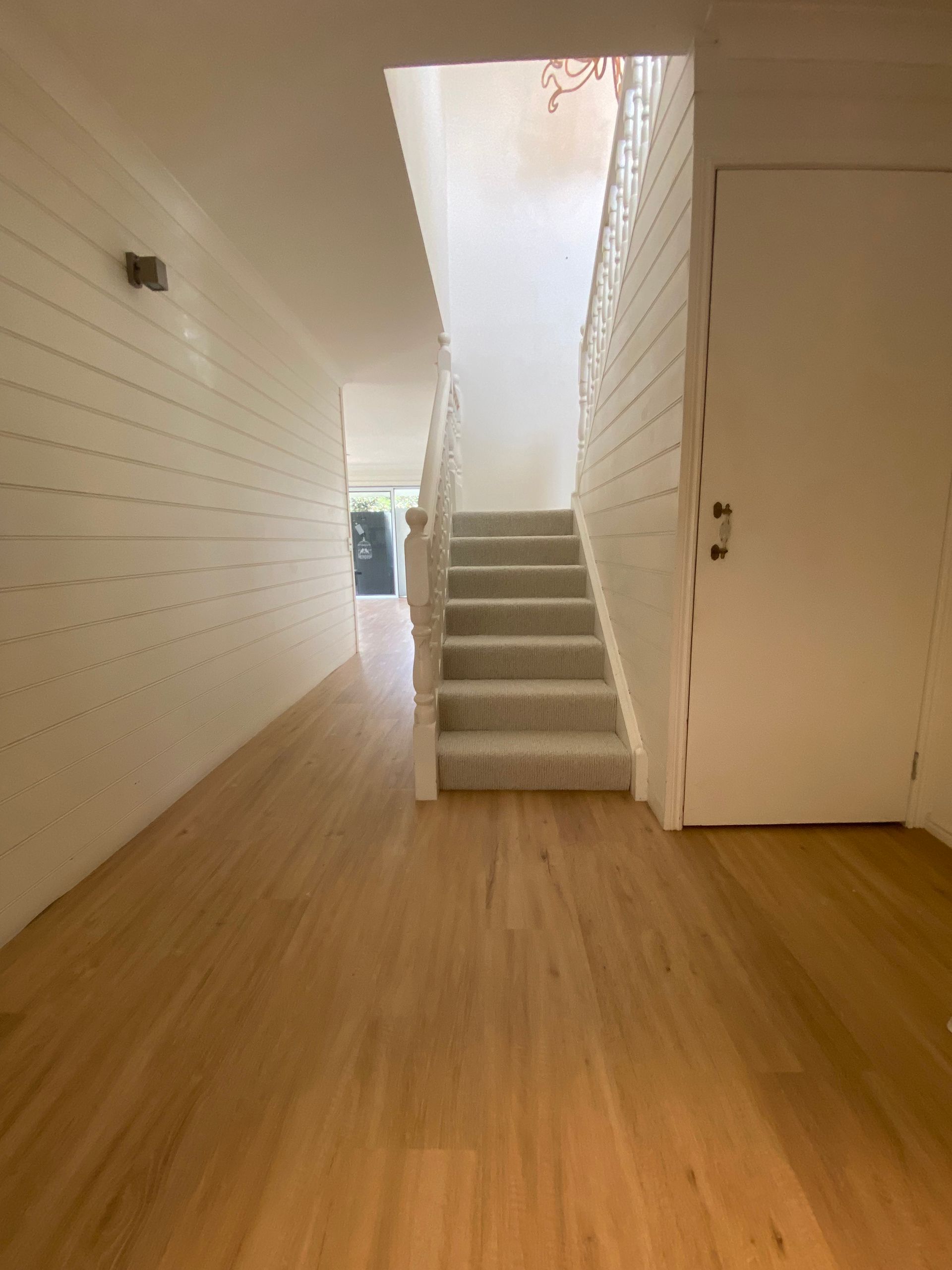Bright hallway with wood floors and stairs leading up to a landing, white walls, and a doorway on the right.