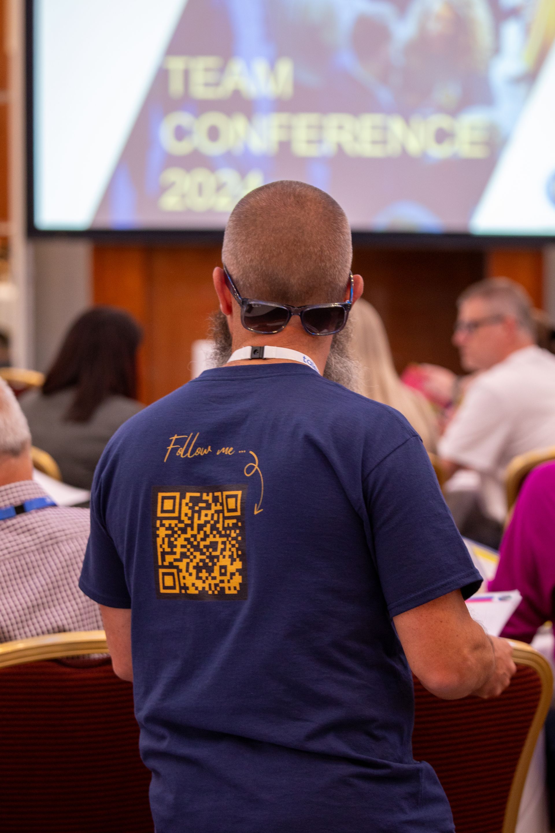 A man wearing a blue shirt with a qr code on the back is sitting in a conference room.