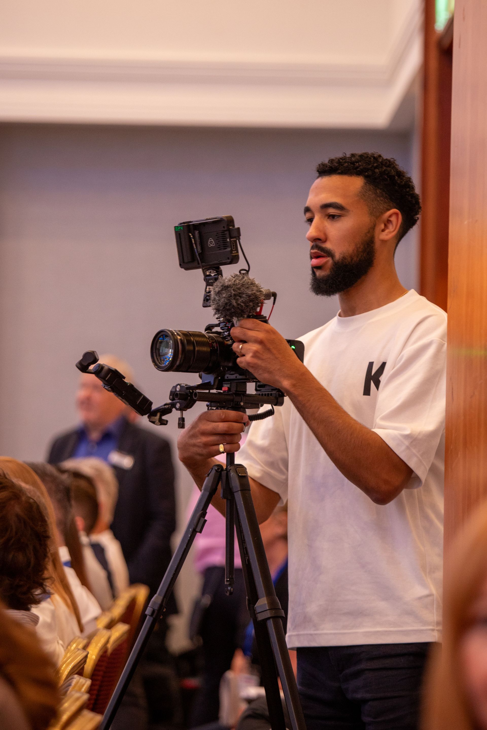 A man is holding a camera on a tripod in front of a crowd of people.