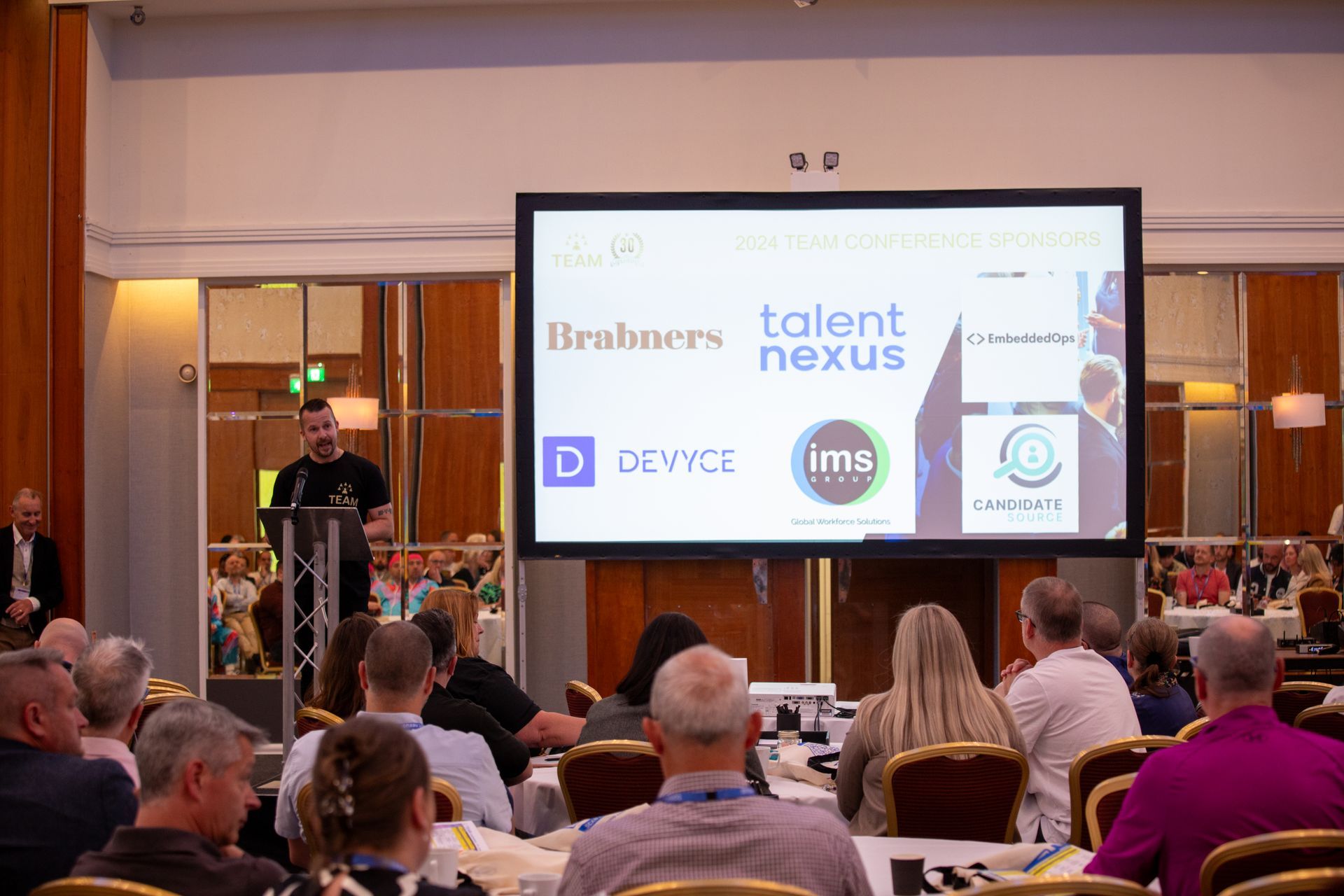A group of people are sitting at tables in front of a large screen.