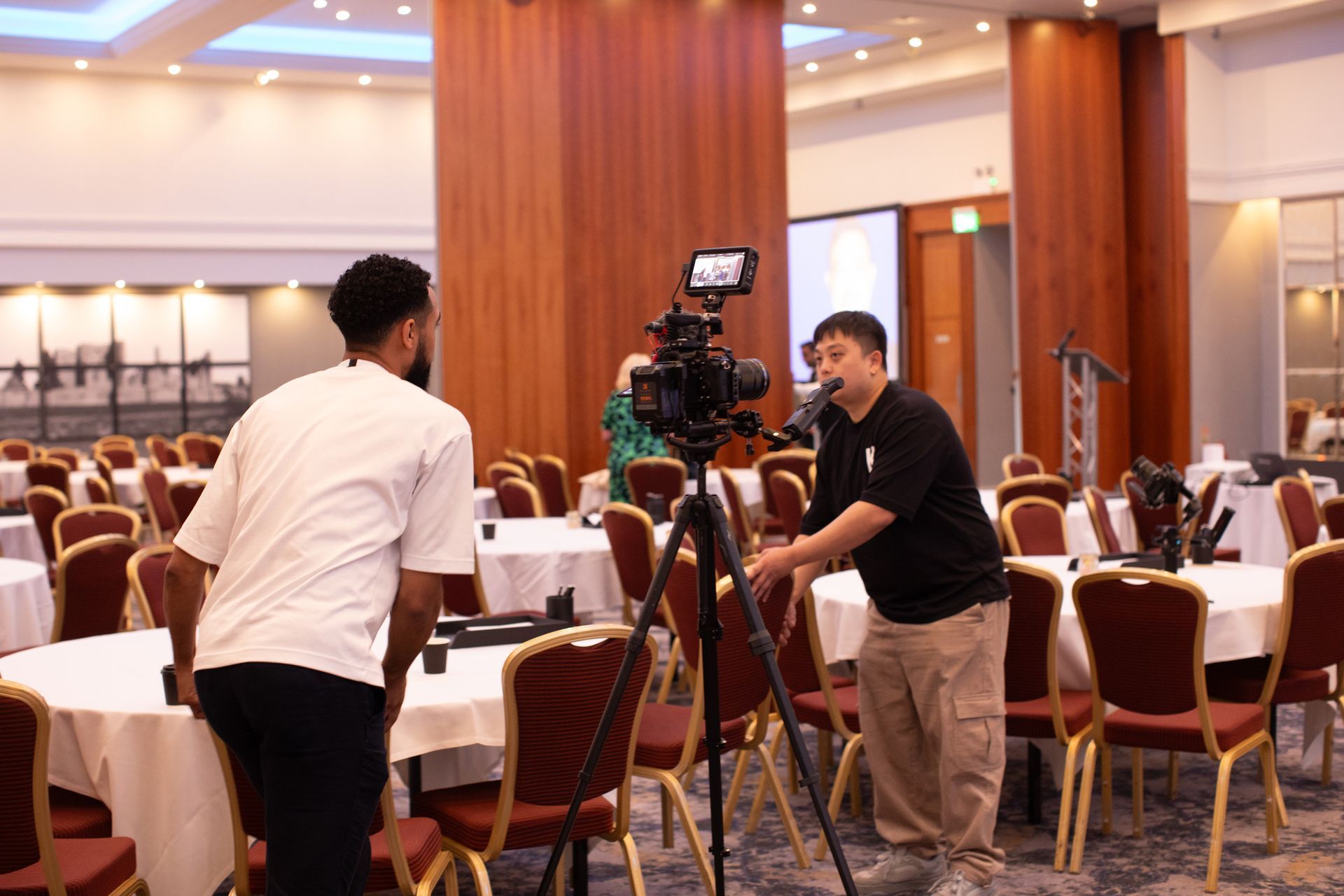 Two men are standing in a room with tables and chairs and a camera on a tripod.