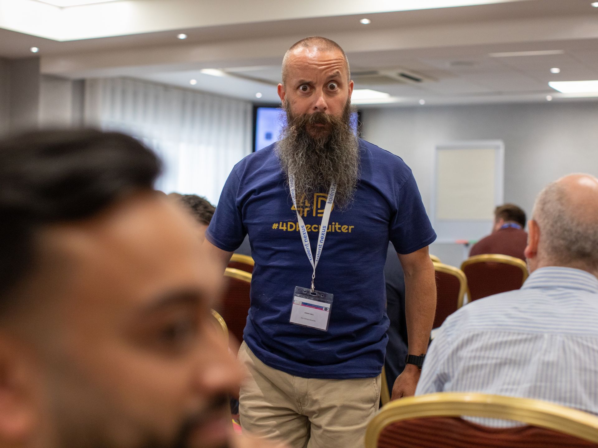A man with a beard is standing in front of a group of people in a conference room.