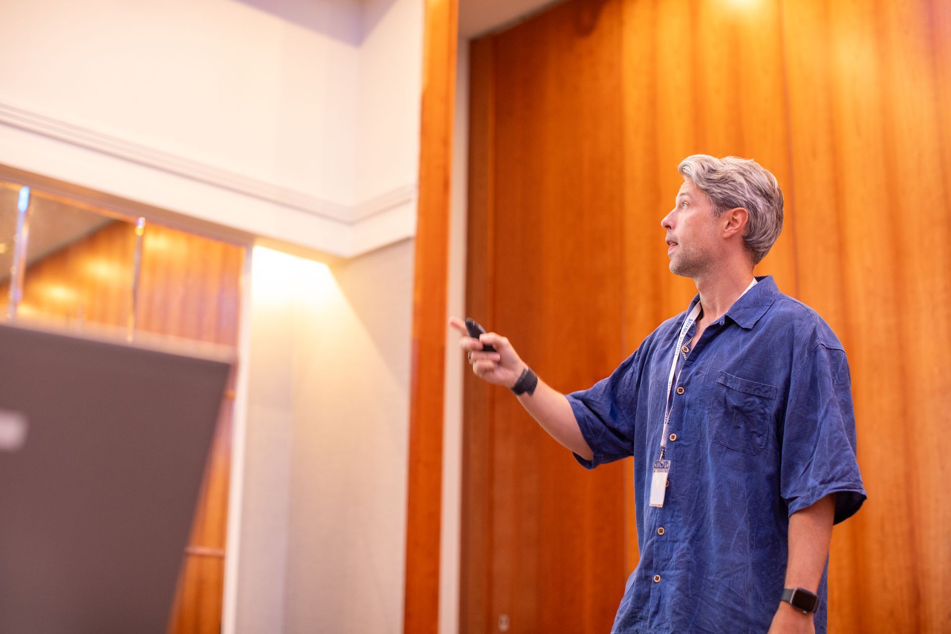 A man in a blue shirt is standing in front of a wooden wall holding a remote control.