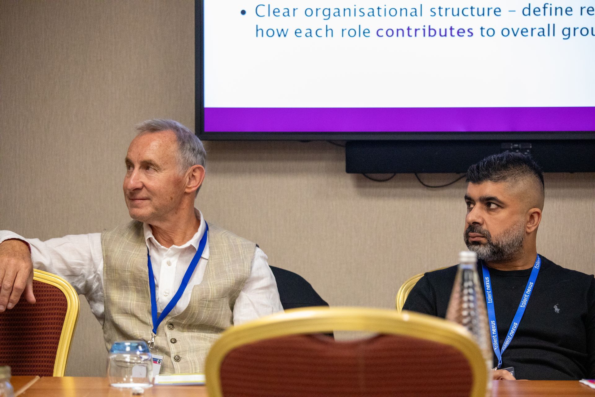 Two men are sitting at a table in front of a large screen.