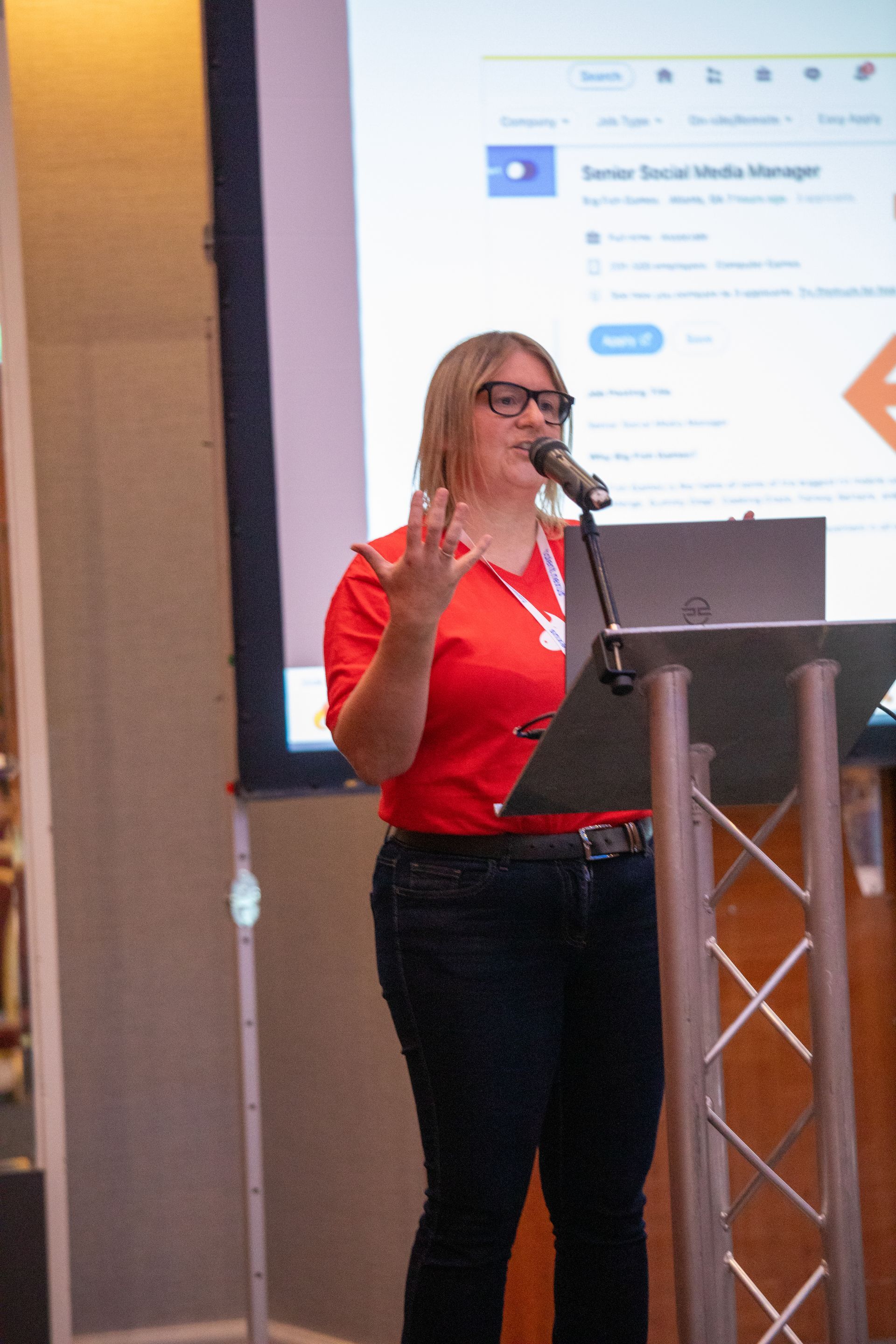 A woman in a red shirt is standing at a podium speaking into a microphone.