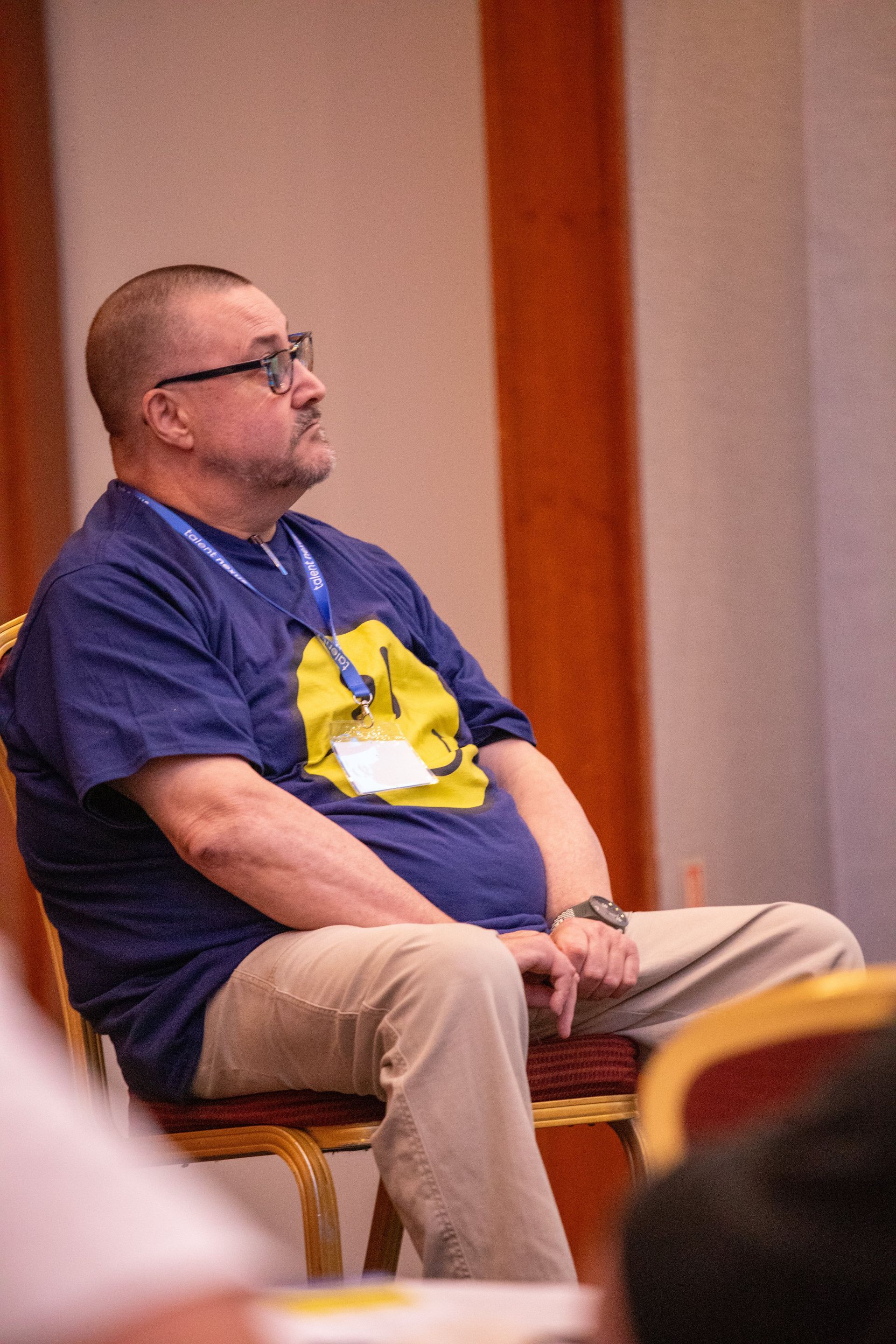 A man wearing glasses and a name tag is sitting in a chair.