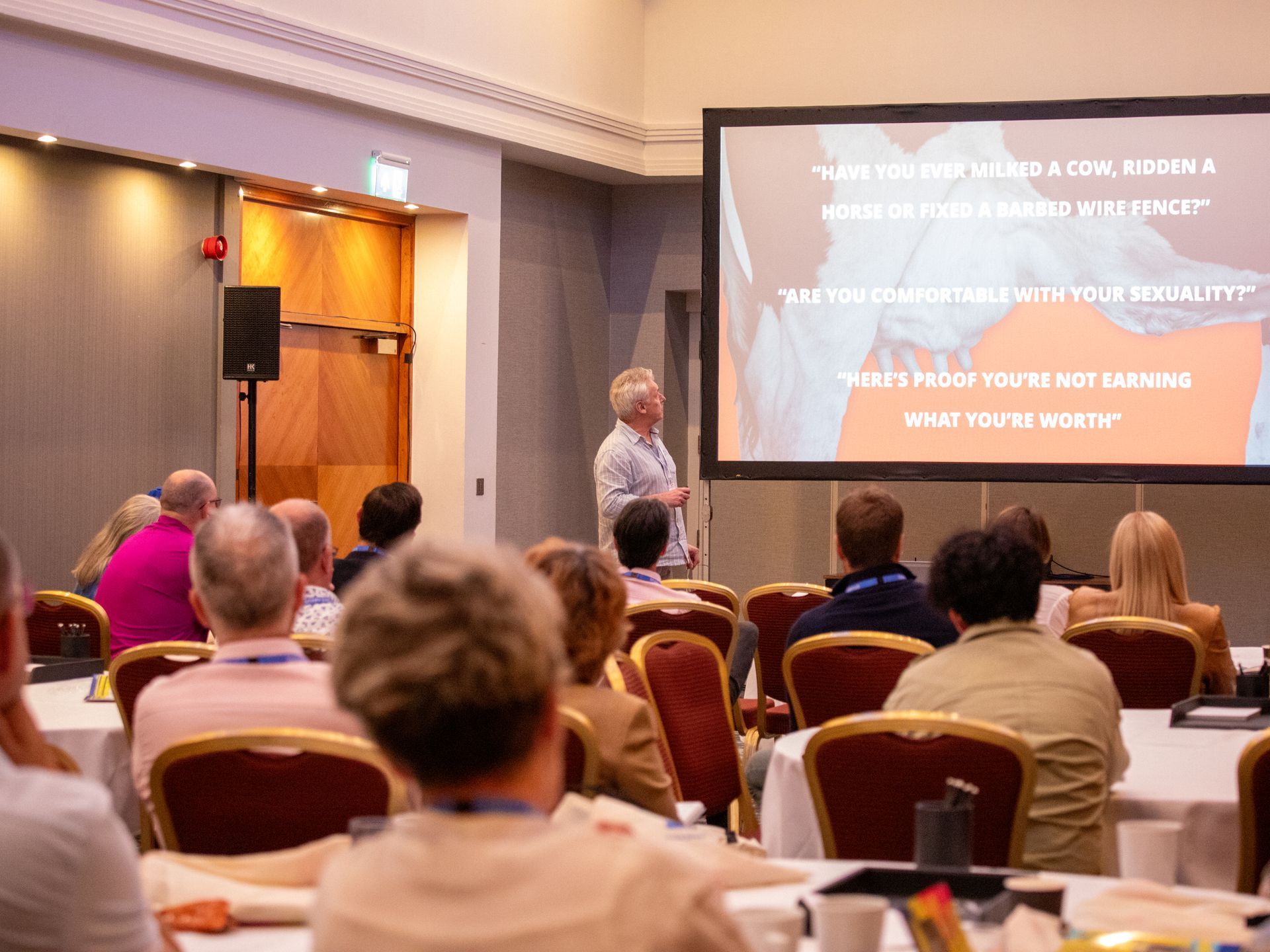 A group of people are sitting at tables in front of a projector screen.