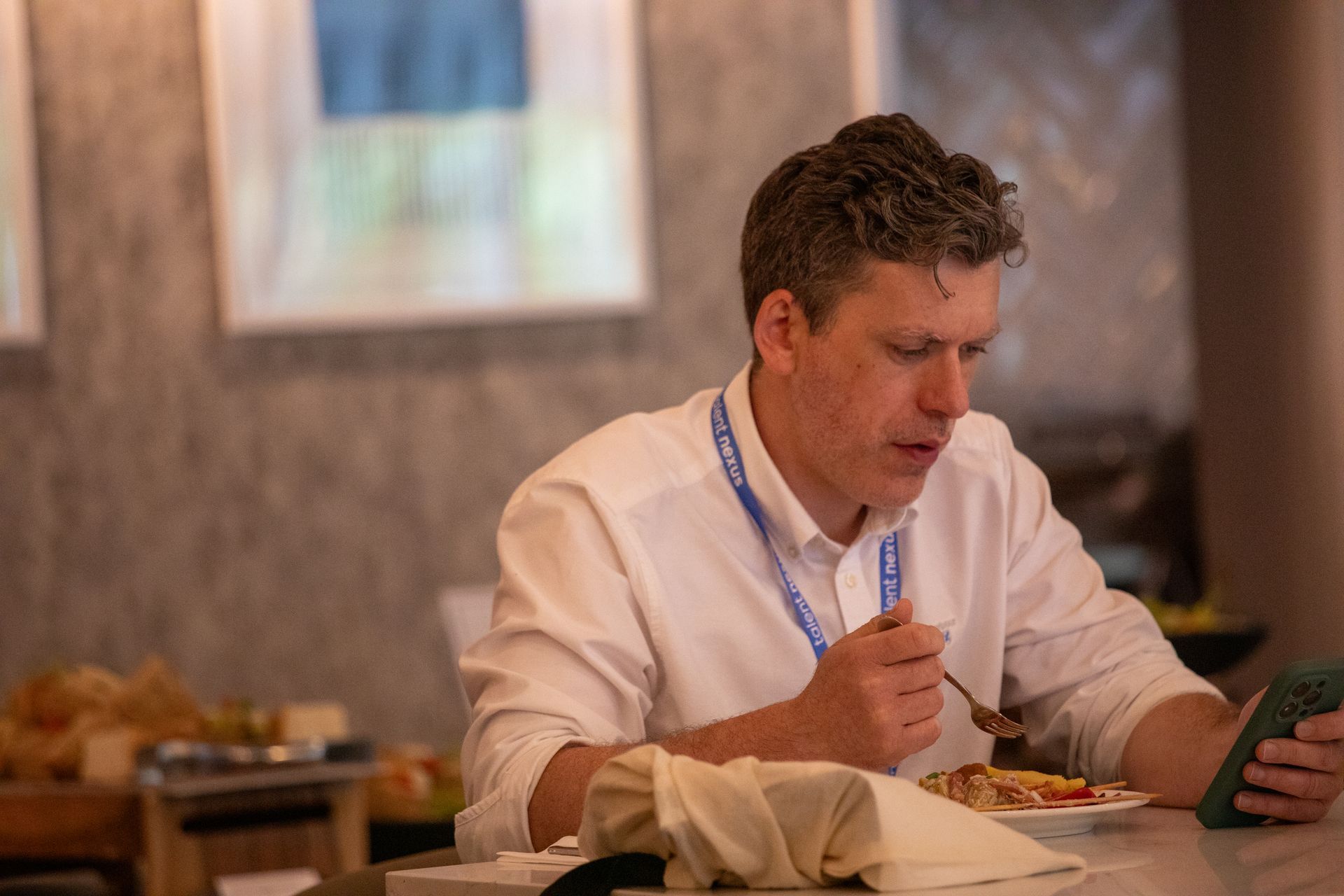 A man is sitting at a table eating food and looking at his phone.