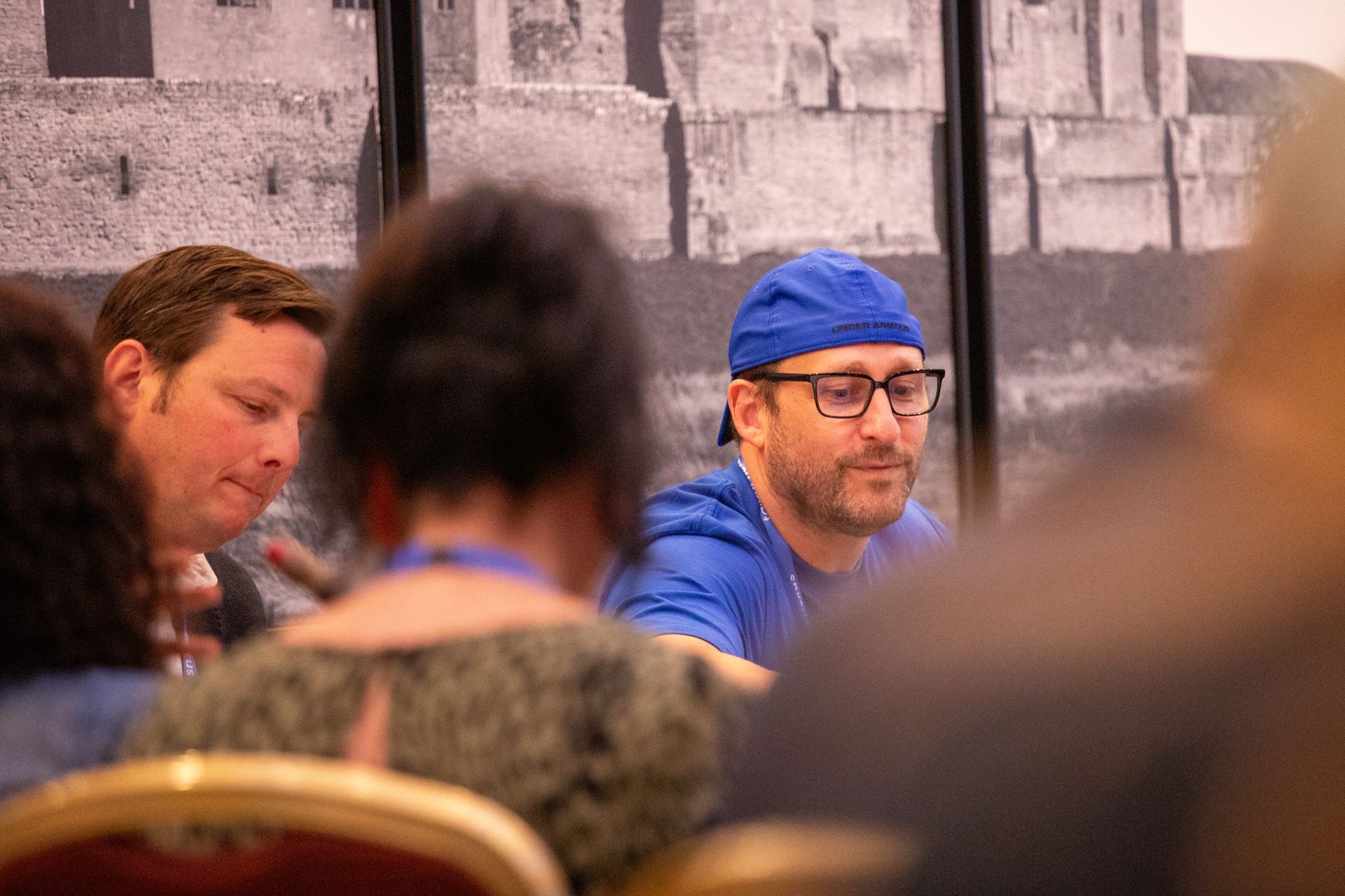 A group of people are sitting at a table with a man wearing a blue hat and glasses.