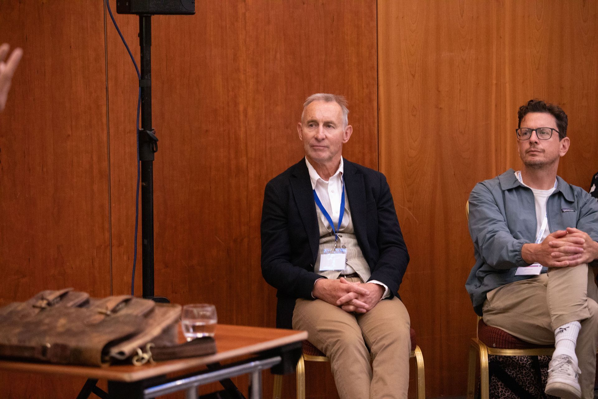 Two men are sitting in chairs in front of a wooden wall.