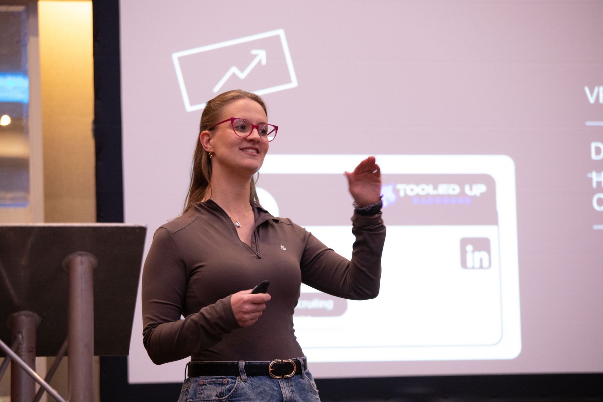 A woman wearing glasses is giving a presentation in front of a large screen.