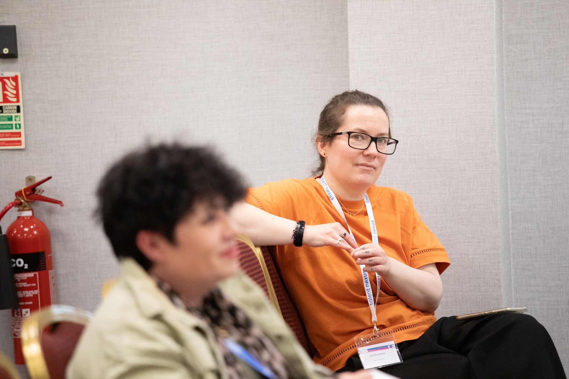 Two women are sitting in chairs in front of a fire extinguisher.