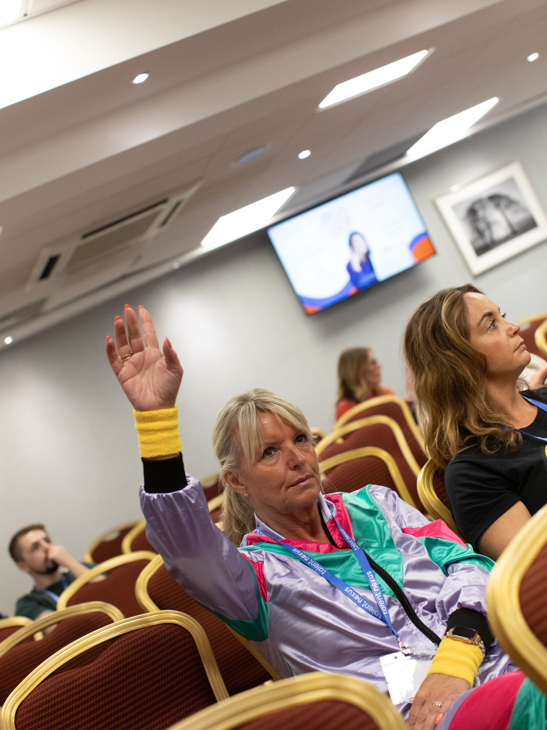 A woman in a purple jacket is raising her hand in a conference room
