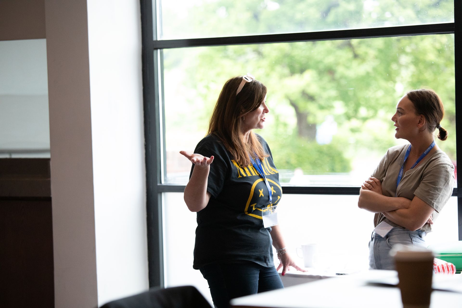 Two women are standing in front of a window talking to each other.
