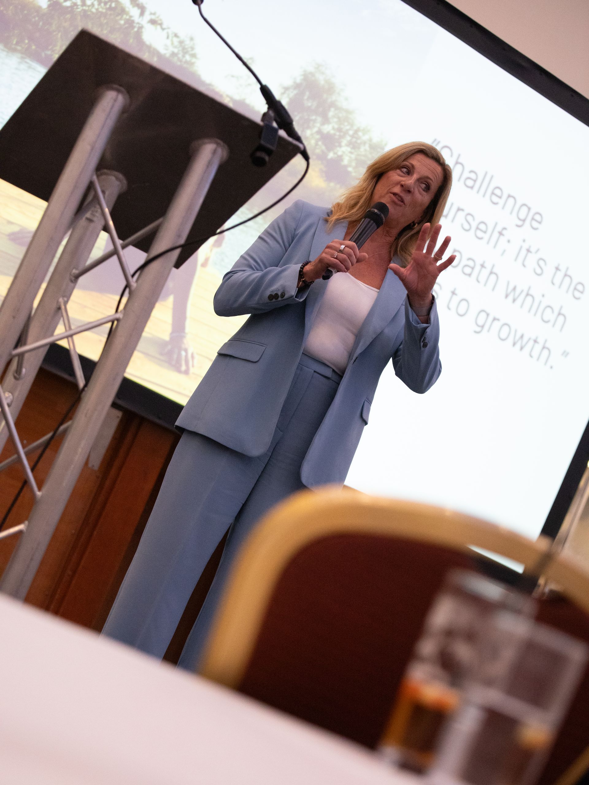A woman in a blue suit stands at a podium speaking into a microphone