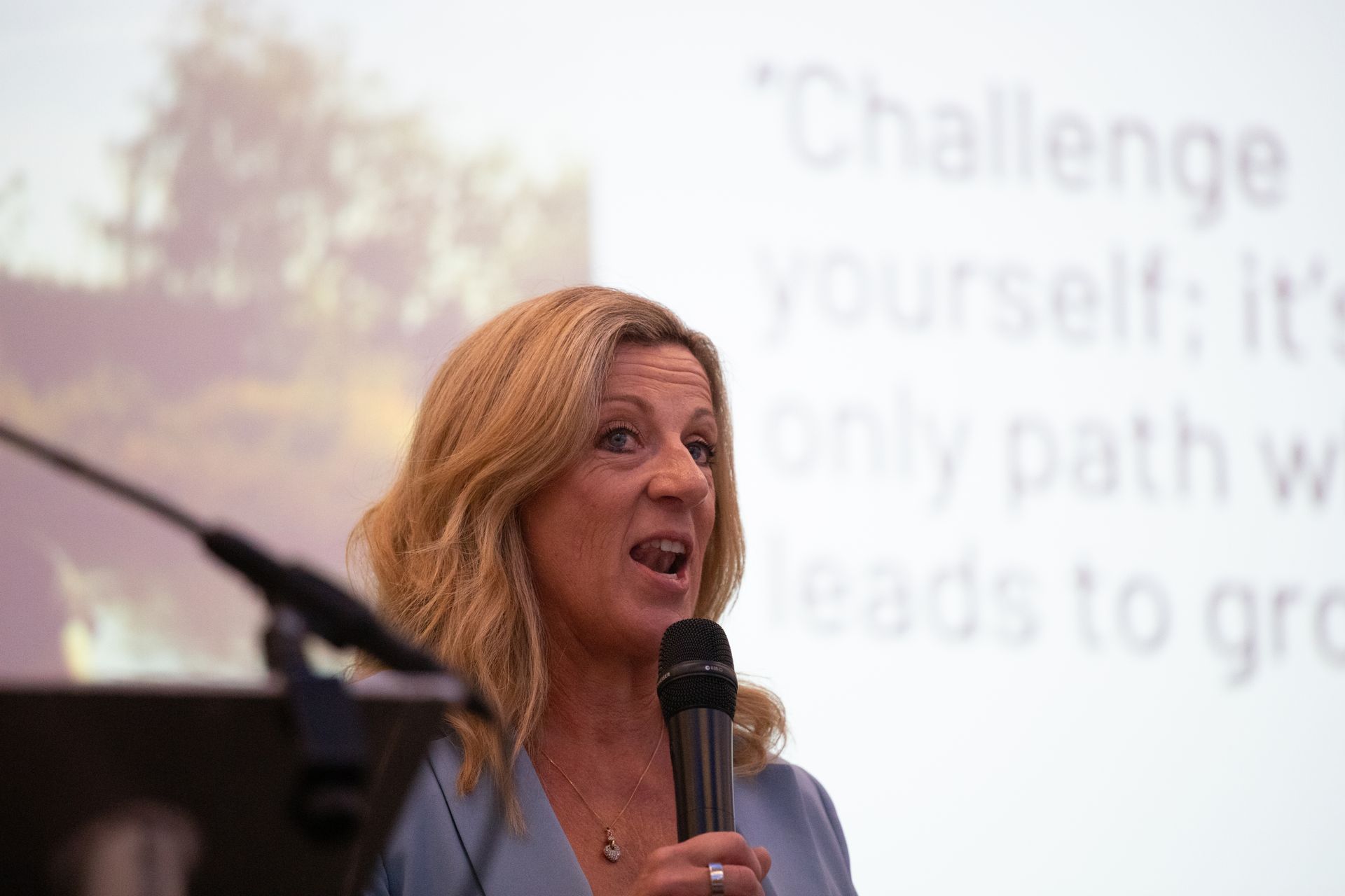 A woman is speaking into a microphone in front of a projection screen.
