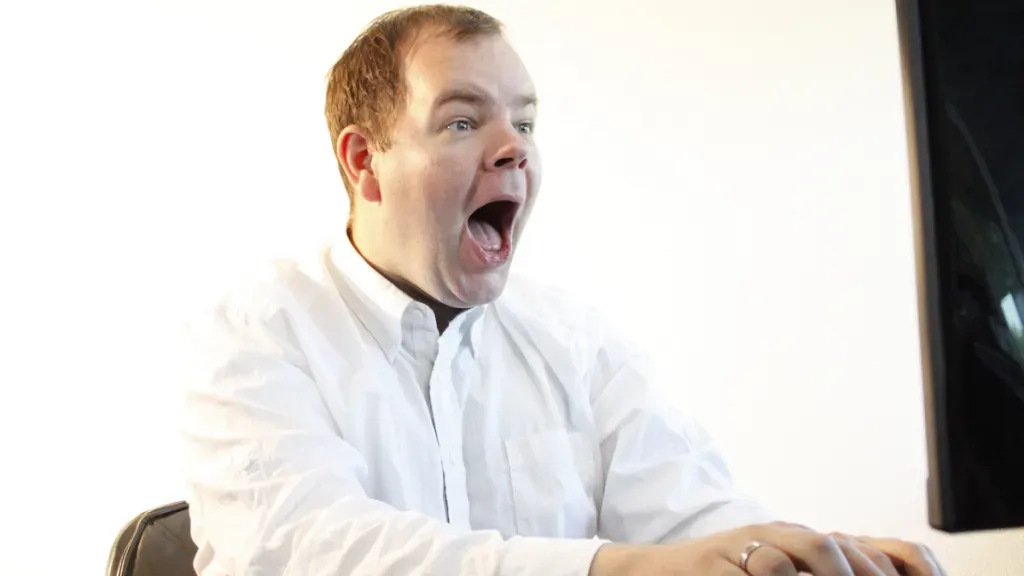 A man in a white shirt is yawning while using a computer.