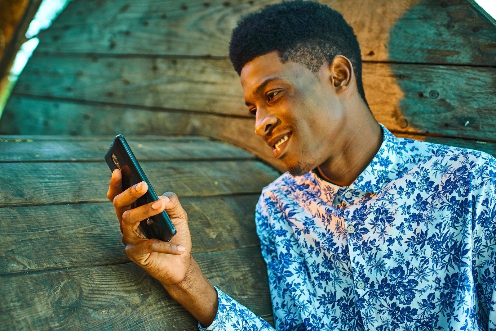 A young man is leaning against a wooden wall while looking at his cell phone.