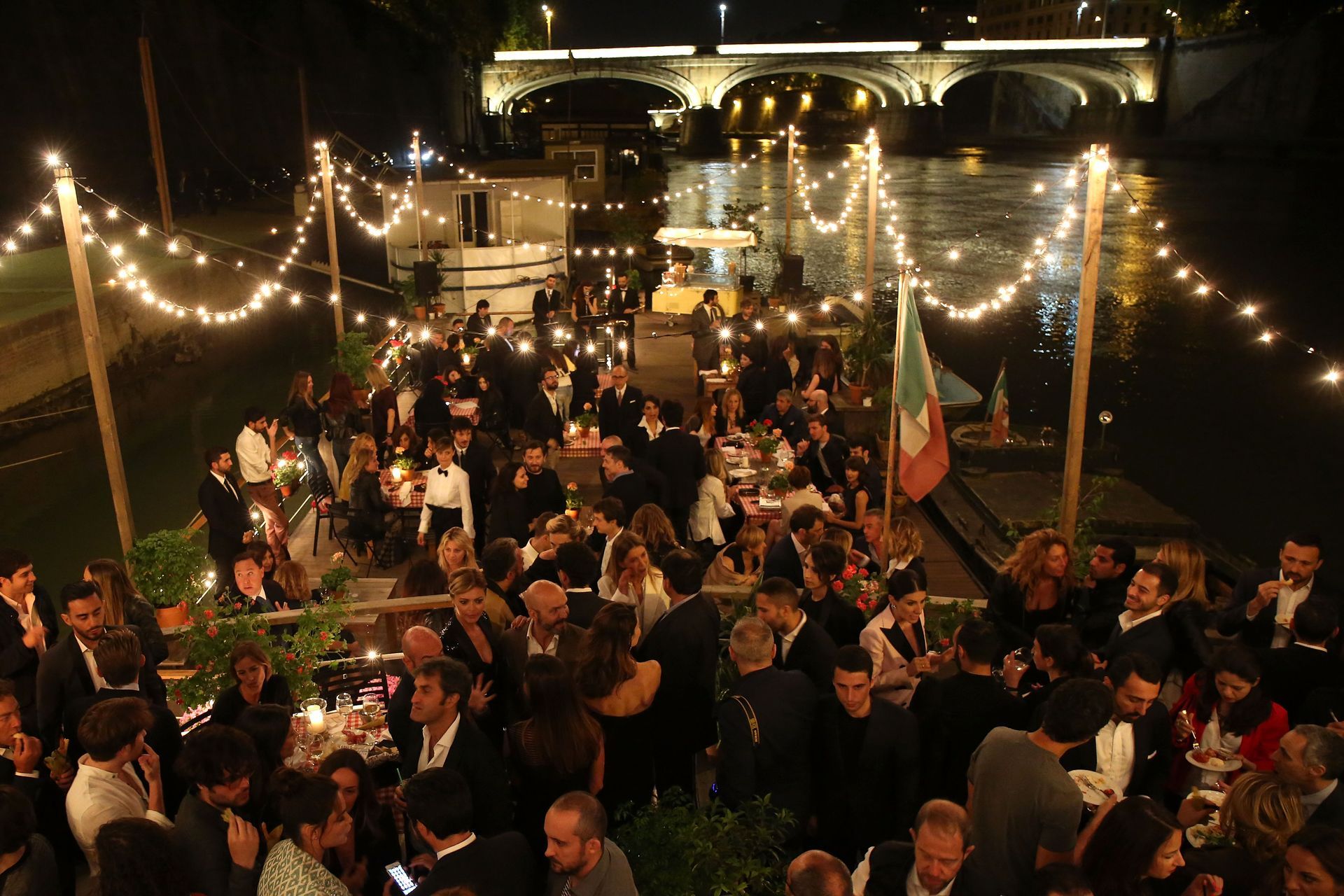 Un folto gruppo di persone è seduto a dei tavoli di fronte a un fiume di notte.