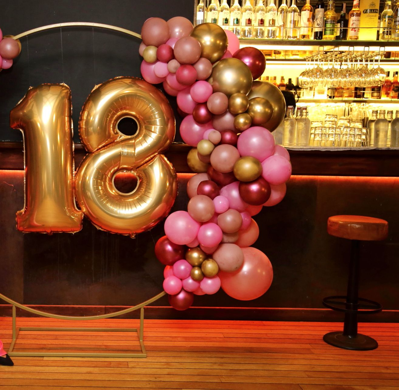 Una donna con un vestito rosa è in piedi davanti a una mostra di palloncini per il suo 18° compleanno.