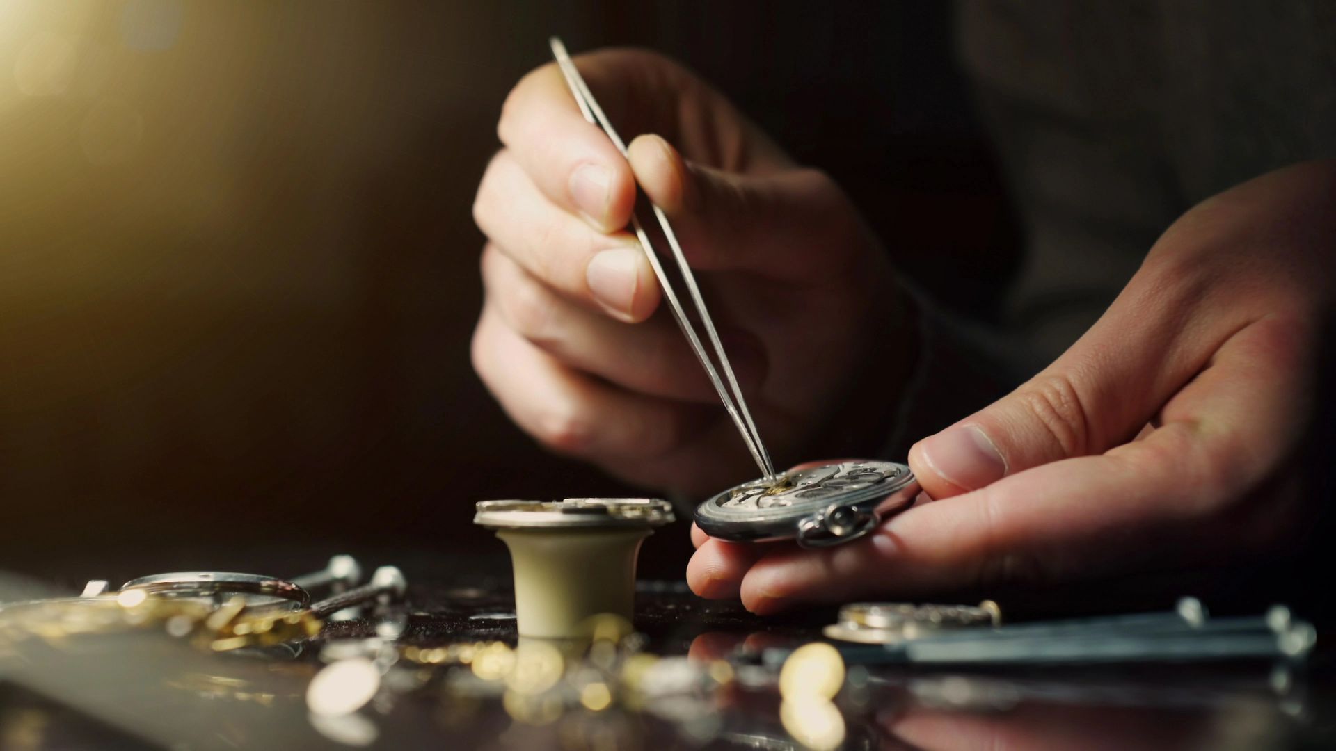 Mani che usano le pinzette per lavorare su un orologio con strumenti, parti e sfondo scuro.