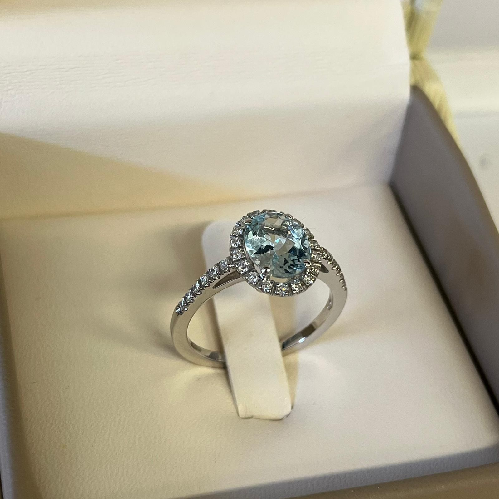 Anello in argento con pietra preziosa ovale blu e alone di diamanti, presentato in una scatola per anelli bianca.