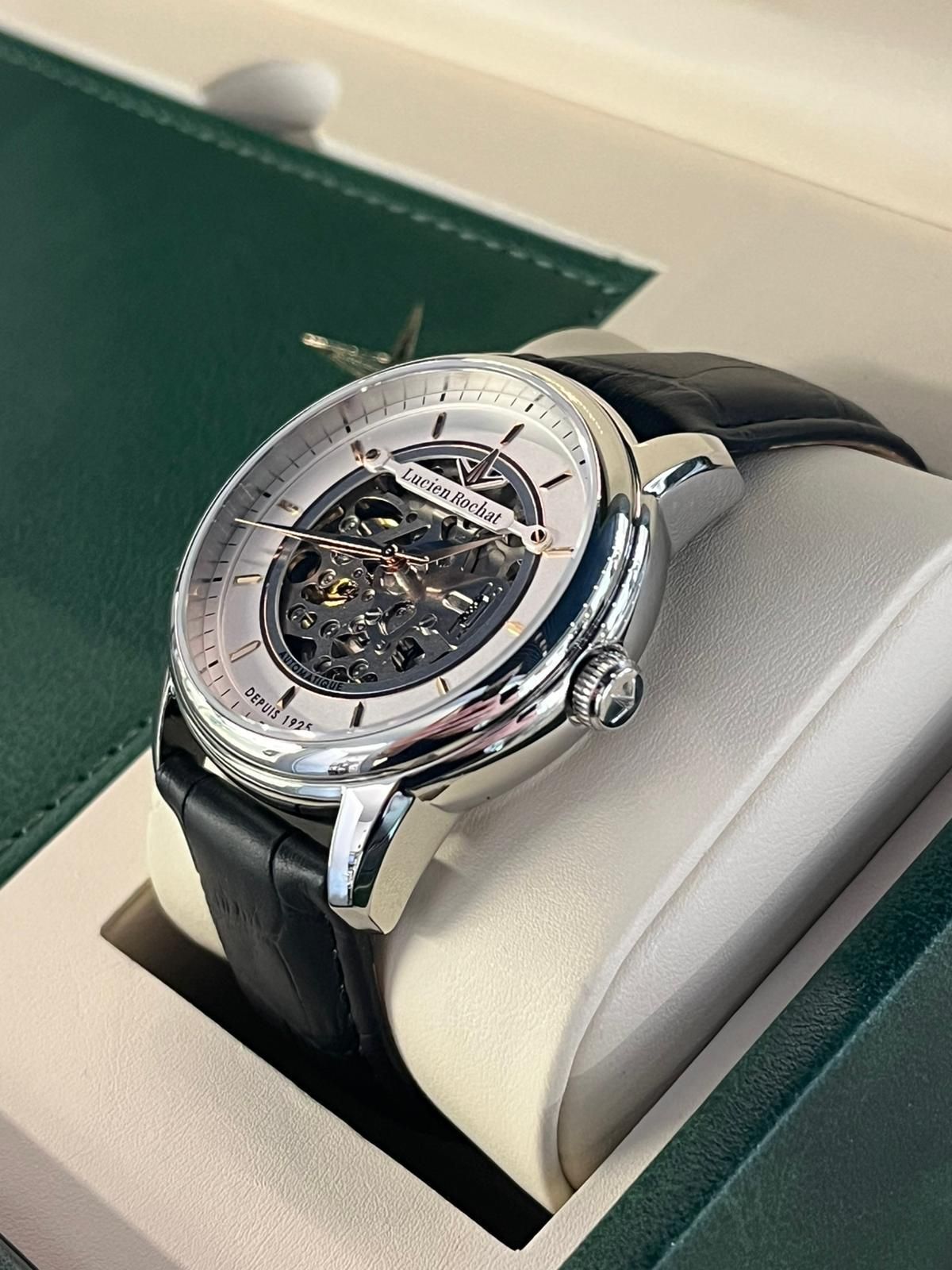 Orologio scheletrato con cassa in argento e cinturino in pelle nera, appoggiato su un cuscino bianco all'interno di una scatola verde.