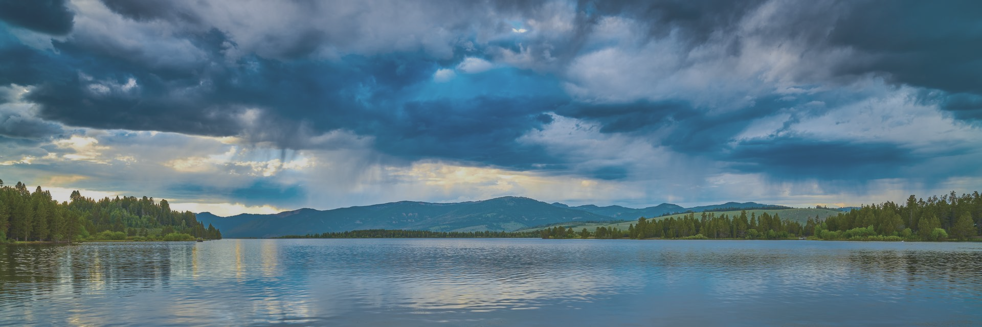 Dramatic cloudy sky over a calm lake with forested shorelines and distant blue mountains