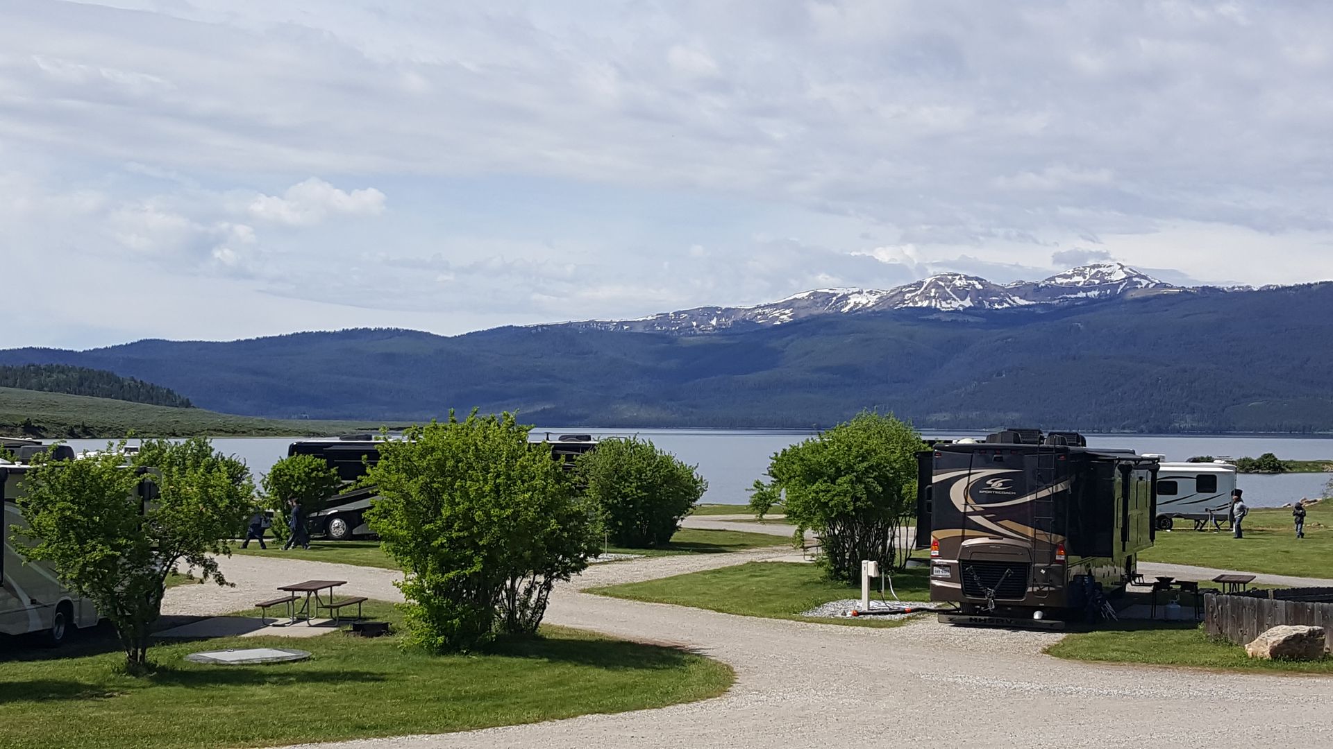 Campground with RV, picnic tables, green trees, and a lake backed by snow-capped mountains under a cloudy sky