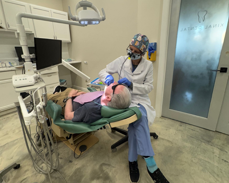 Dentist examining a patient in a dental office. The dentist is wearing a mask and scrubs. The patient is in a chair.