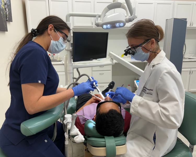 Two dental professionals wearing masks and gloves working on a patient in a dentist's chair.