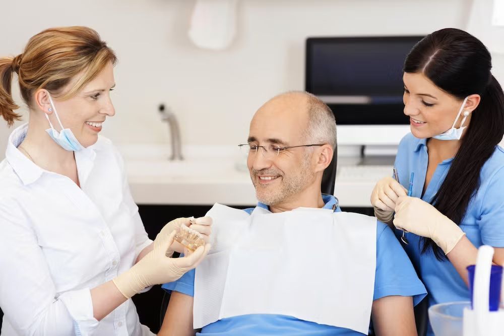 Dentist and assistant with a patient; dentist holding dental mold, all smiling in an office setting.