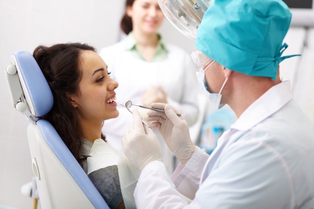 Woman in dental chair, mouth open, dentist examining teeth, assistant in background.