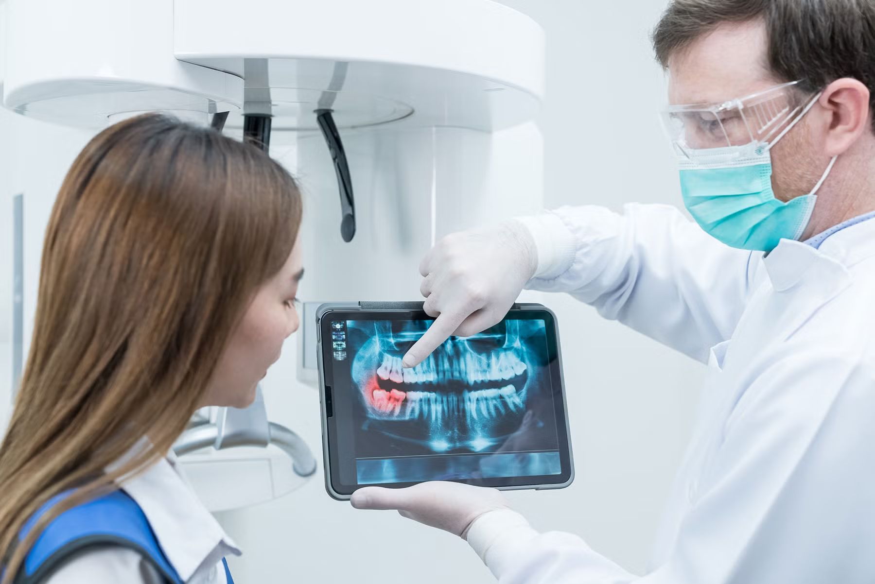 Dentist shows patient a dental X-ray on a tablet, pointing to a tooth. Dental office setting.