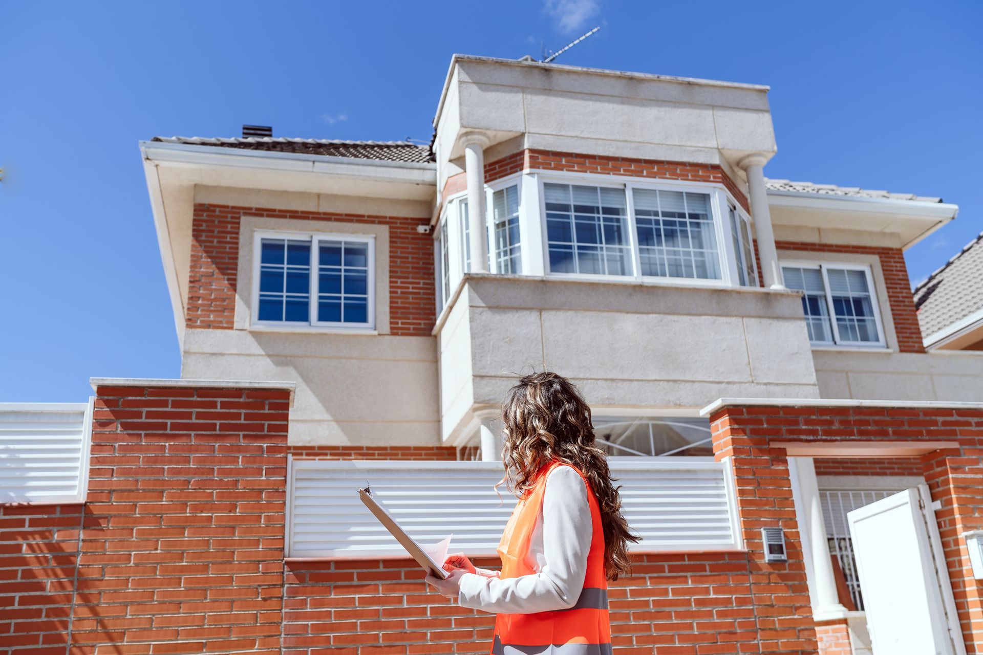 A woman is standing in front of a brick house holding a tablet.